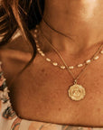 A close-up image of a person wearing a gold-filled necklace with a Medallion design, layered with a pearl chain.