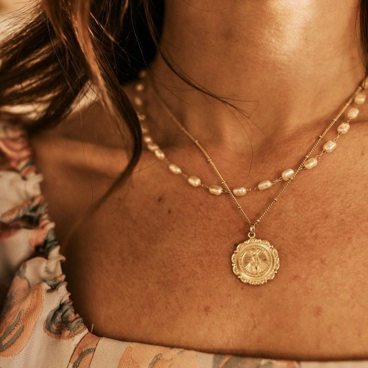 A close-up image of a person wearing a gold-filled necklace with a Medallion design, layered with a pearl chain.